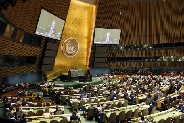 UN assembly hall with people seated watching a speaker on screen
