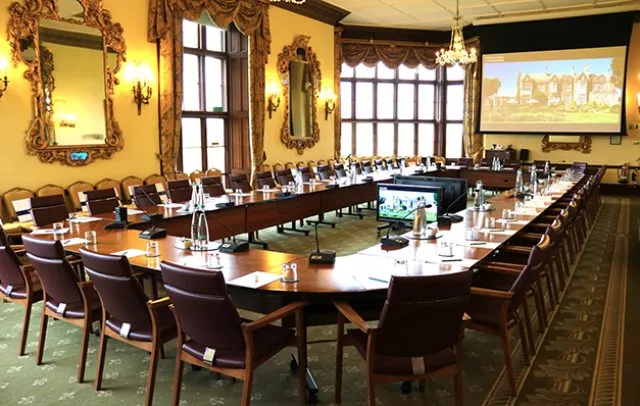 Wilton Park's conference room with a large conference table, chairs, a projector screen and a monitor
