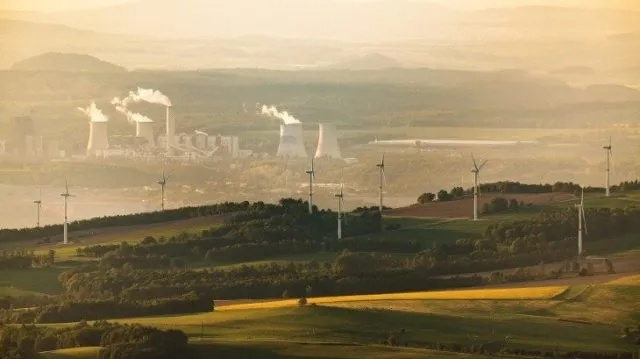 Faint view of chimneys with smoke in the background, with a more vibrant view of a wind farm in the foreground
