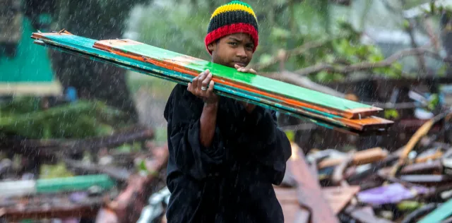 A child carries planks of wood over their shoulder, as they walk through the rain