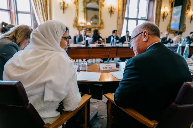 Two participants at a Wilton Park dialogue smile at one another as they speak at the conference table. Other participants are visible in the background