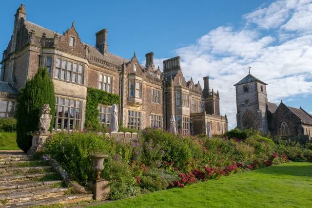 An external shot of Wiston House with the low autumn sun shining on it. The House is visible in the centre of the shot, with flowers in the foreground and a church in the background.