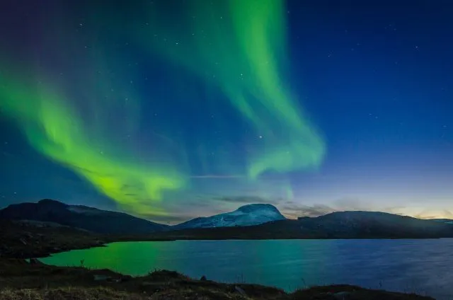 A display of blue and green Northern Lights in the sky over a lake and mountains