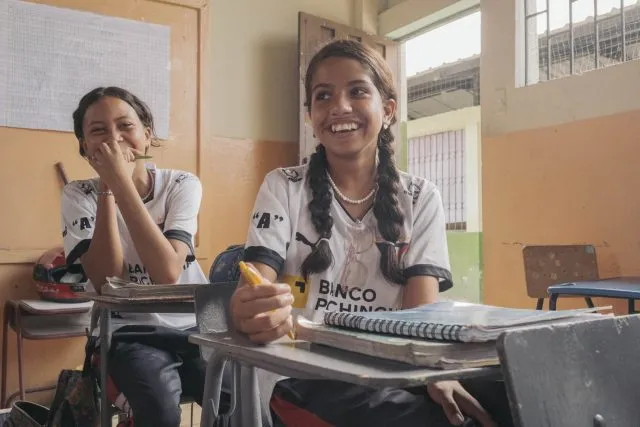 Two young girls sit at desks in an Ecuadorian classroom, smiling.