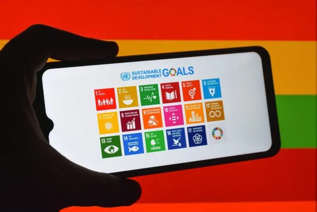 A hand holds a mobile phone. On the screen are coloured blocks showing the sustainable development goals