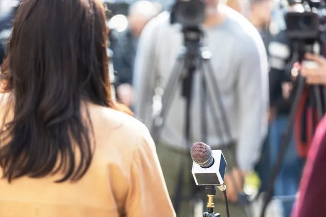 A woman stands by a microphone, facing away from the camera. A cameraman is visible filming her in the background.