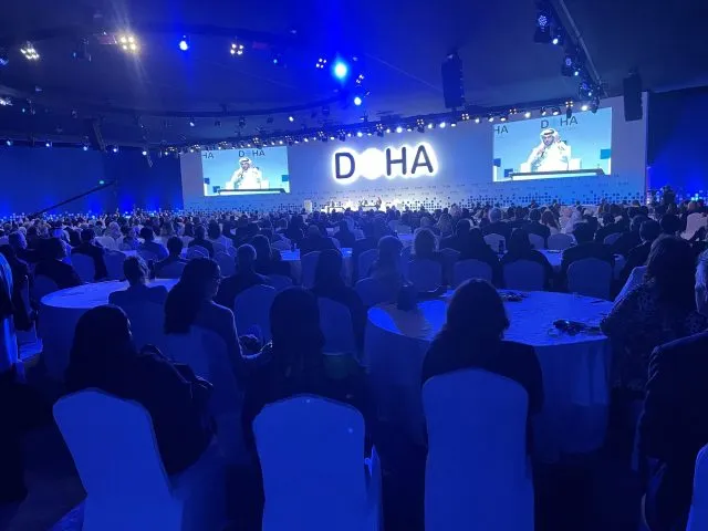 Participants at the Doha Forum are seated in a large, darkened hall. The word 'DOHA' is displayed on a large screen on a stage.